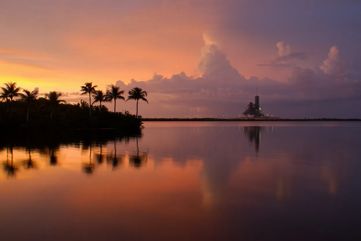 Kennedy Space Center en Floride au crépuscule avec le pas de tir 39B au loin, palmiers en premier plan et ciel orangé se reflétant sur l'eau