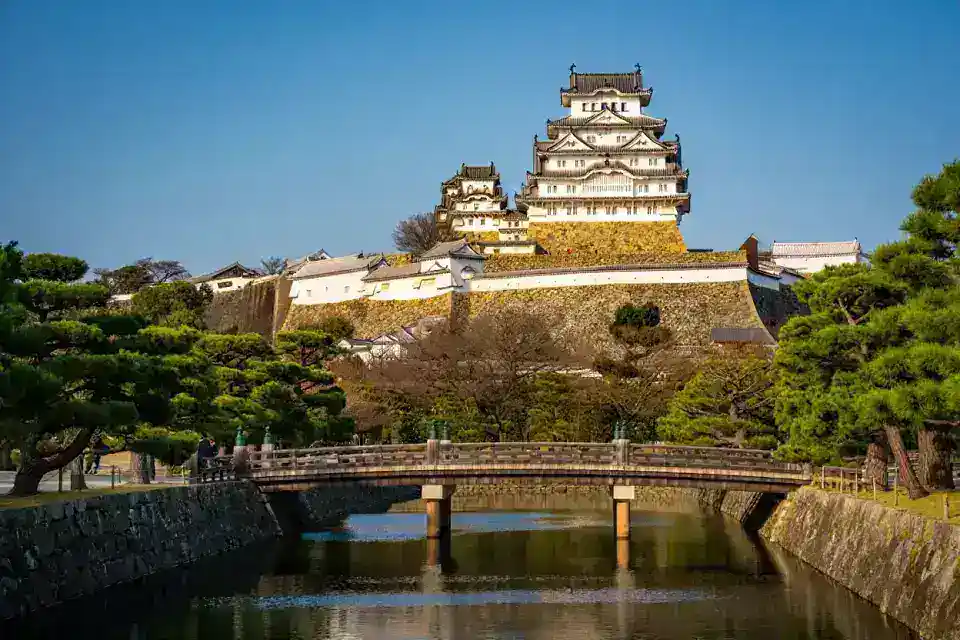 Château d'Himeji vu depuis les jardins japonais, murs blancs à la chaux dominant un pont de pierre au-dessus des douves, lumière dorée d'après-midi