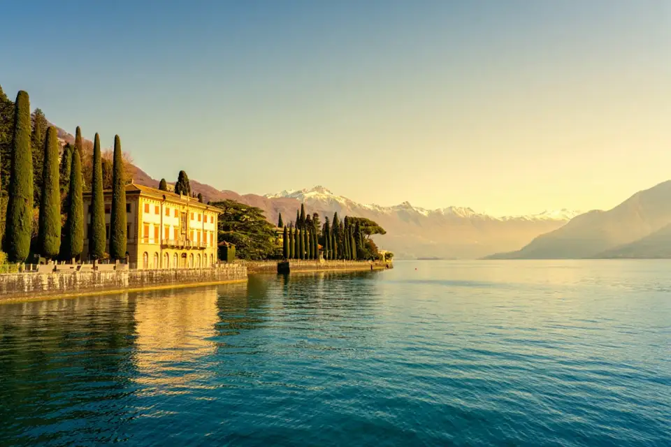 Vue panoramique du lac de Côme depuis la rive ouest à Cadenabbia, avec un palazzo historique au bord de l'eau, des cyprès et les Alpes enneigées en arrière-plan