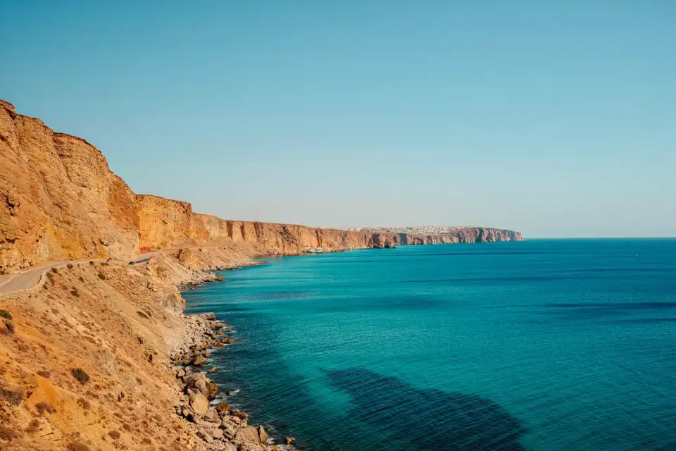Route côtière méditerranéenne au Maroc, falaises ocre plongeant dans une mer turquoise, route étroite longeant la côte du Rif, lumière dorée du matin